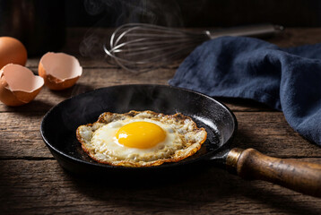 Close-up of a perfectly fried egg with a bright yellow yolk and crispy edges, steaming hot in a rustic cast iron pan on a dark wooden table, with f...
