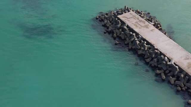 Drone footage captures a concrete breakwater surrounded by dark tetrapods in turquoise coastal water, showing calm waves, shoreline details and peaceful marine atmosphere.