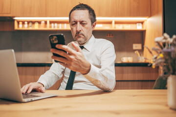 Businessman working on laptop and smartphone in a home office, multitasking between devices while focusing on digital tasks.