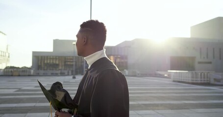 African American man in graduation gown at sunset on plaza holding cap inspecting yellow tassel
