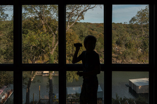 Silhouetted child holding binoculars in front of a window near lake
