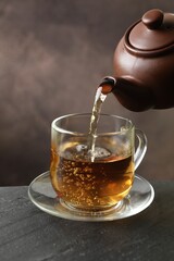 Pouring hot tea into cup from teapot at black table, closeup