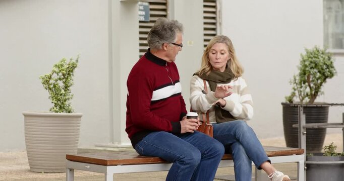 Tapping smartphone Senior couple sitting bench comparing watches walking path holding coffee phone