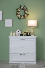Christmas decor and lamp on white dresser near green wall indoors. Interior design