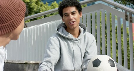 Upon gripping soccer ball Diverse male friends sitting on steps beside blue wall discussing tactics