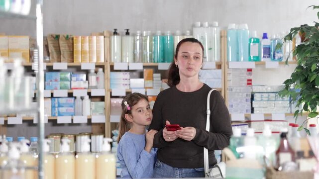 Woman and girl in pharmacy with phone, looking for something in drug store, bored child pulls mother by hand. Parent scrolls screen in search of electronic prescription. 