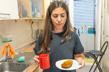 Woman standing in her kitchen holding a red mug and a plate with toast for breakfast
