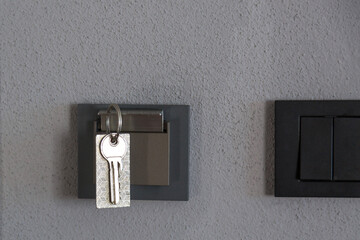 Metallic key with a patterned keychain hanging on a modern gray wall hook near a black light switch, concept of security, home access, safety and minimal interior design