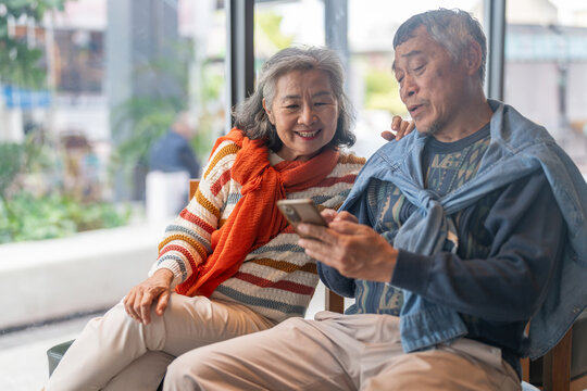 Senior asian couple using smartphone together sitting and smiling at cafe with digital technology, mobile banking, online shopping or telehealth services for elderly, modern lifestyle after retirement - Powered by Adobe