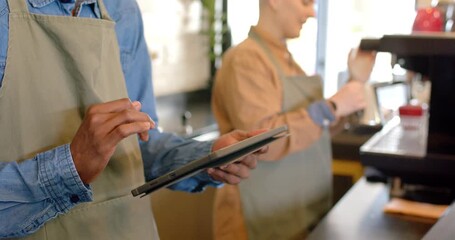 Diverse coworkers reviewing order, tapping tablet, frothing milk with steam wand in pitcher at bar
