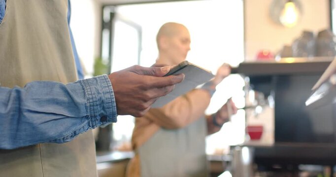 Diverse coworkers tapping tablet viewing order, tamping portafilter and steaming pitcher at counter