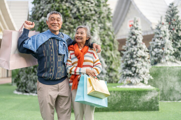 Happy senior asian couple holding shopping bag in front of snowy Christmas tree, enjoying winter holiday season, new year end sale festive shopping, senior lifestyle, love, joy and holiday celebration