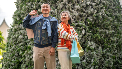 Happy senior asian couple holding shopping bag in front of snowy Christmas tree, enjoying winter...