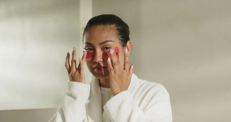 With red under-eye gel patches Asian woman pressing massaging eyes in room to boost circulation