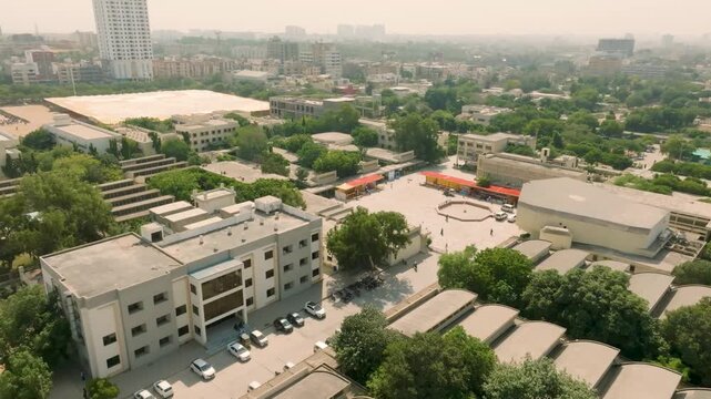 Drone pan over a central NED University campus building and adjacent parking lot filled with cars, with students gathered in the distance and hazy city skyline beyond.