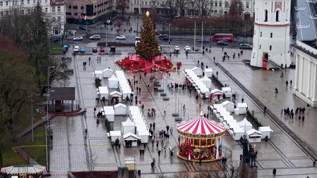 High-angle daytime view of a Christmas market with a decorated tree, carousel and rows of white huts, capturing seasonal shopping, open-air activity and festive atmosphere on a wet plaza.