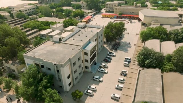 Aerial orbit view of NED University of Engineering and Technology campus parking lot in Karachi, featuring parked cars, students, and campus buildings under a sunny sky.