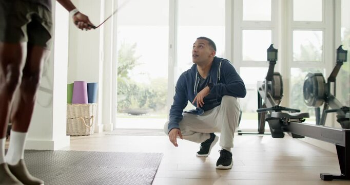 Male coach in home gym seeing trainee skipping rope, crouching, clapping and guiding form near mat
