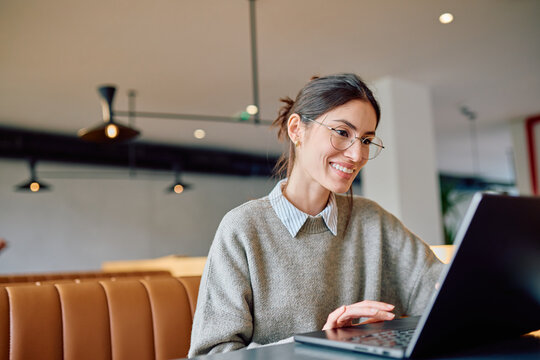 Young smiling woman wearing glasses using laptop at a modern cafe, enjoying remote work and online learning experience