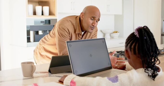 Mature father leaning over laptop guiding African American daughter typing schoolwork on counter