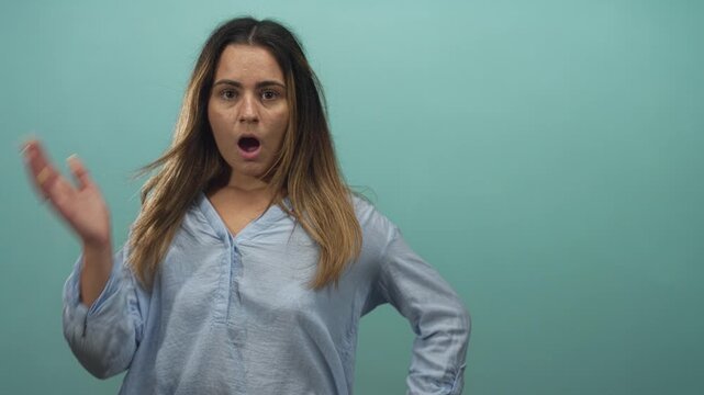 Woman mouth open hand raised in mint green studio showing open mouth and expressive gesture, wearing light blue blouse with loose sleeves; surprise reaction.