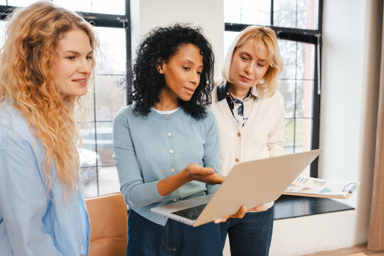 Diverse women collaborating on laptop, discussing business strategy during office meeting - Powered by Adobe