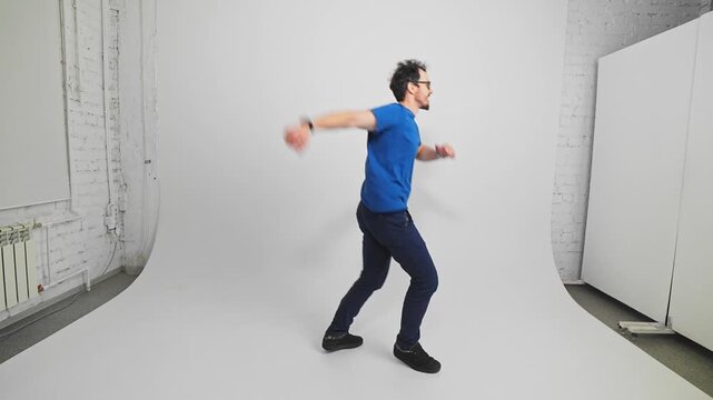 Side view of cheerful Caucasian guy in T-shirt and trousers jumping and moving filmed in slow motion. Young adult bearded guy hops showcasing fun and energy in studio with white walls. Movement theme.