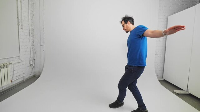 Caucasian bearded man is stepping on diagonal line holding hands by side like equilibrist filmed in slow motion in white studio. Young adult guy is moving carefully on narrow way trying not to fall
