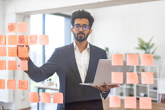 An Indian businessman uses sticky notes and a laptop to plan a project in an office setting - Powered by Adobe