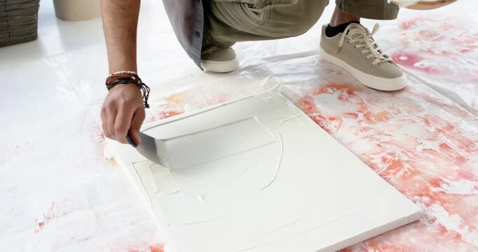 Upon dipping putty knife into white medium man is spreading it onto canvas for textured base