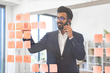 An Arab businessman smiles while talking on the phone and looking at sticky notes on a glass wall