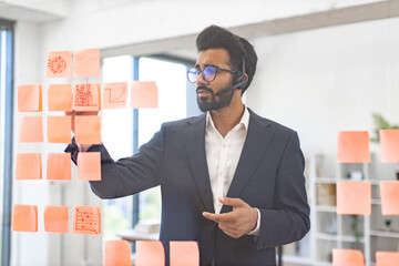 An Indian businessman wearing a headset gestures towards sticky notes during a presentation