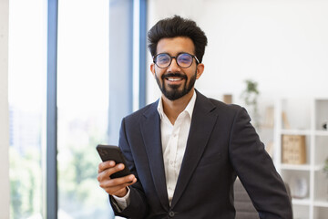 An Indian businessman smiles while holding his phone in a modern office setting