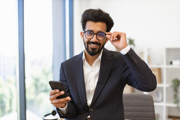 An Indian businessman in a suit smiles while holding his phone and adjusting his glasses
