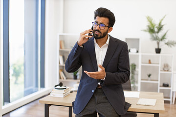 An Arab businessman in a suit gestures while speaking on his phone in a modern office setting