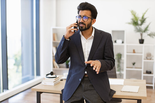 An Indian businessman wearing glasses looks concerned while speaking on his phone in an office setting