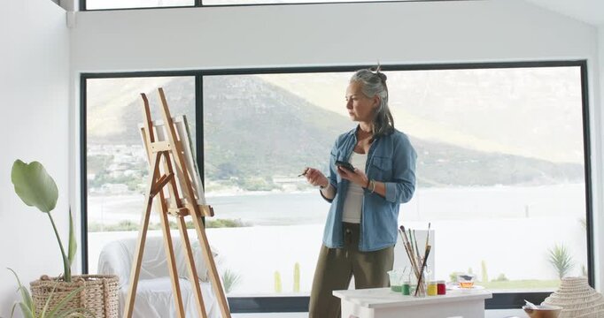 Mature female artist holding brush, gazing at canvas, scrolling smartphone for reference in studio