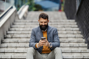 Portrait of smiling businessman sitting on stairs with cellphone in hands and texting messages at financial district downtown.