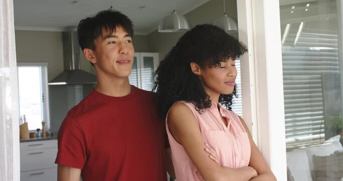 Diverse couple gazing out window, man entering wrapping waist affectionately laughing in kitchen