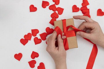 On a white surface, someone is tying a red ribbon around a small, brown gift box. Many small red hearts are scattered around the box, suggesting a Valentine's Day theme.