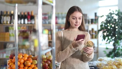 Attractive long-haired woman choosing beer in grocery shop, scanning QR code on can using smartphone to add items to digital cart and process online payment. High quality 4k footage - Powered by Adobe