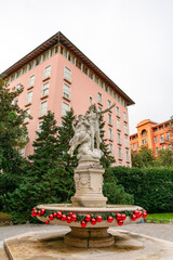 Obraz premium Historic stone fountain in Opatija adorned with bright red Christmas ornaments and festive greenery, surrounded by pastel buildings and evergreen trees