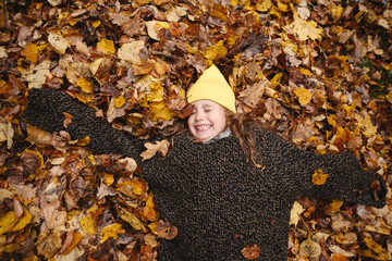 Smiling child in yellow hat lying in autumn leaves at park