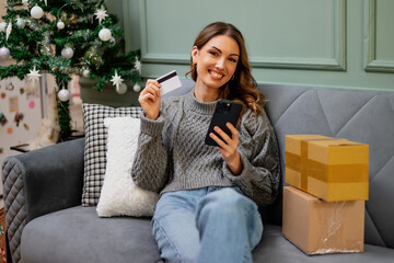 A smiling woman sits comfortably on a sofa, holding a credit card and smartphone, with delivered packages beside her and a decorated Christmas tree in the background.
