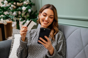 Sitting comfortably on a sofa, woman shows her credit card while using her smartphone for online shopping.