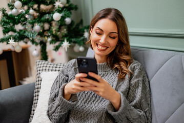 Young happy smiling woman is sitting on couch in her home and using smartphone. Decorated Christmas tree in background.