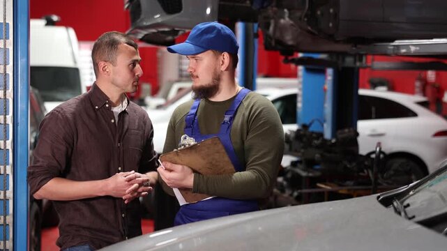 Positive young auto-mechanic in blue overalls communicating with male client of car workshop about service fees indicated on sheets