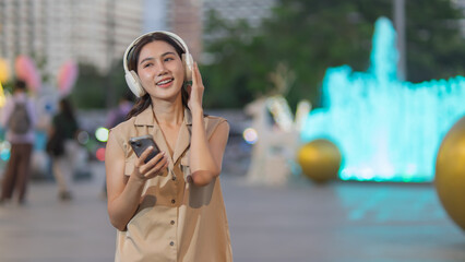 A young woman enjoys music in the city, smiling with her smartphone and wireless headphones. Stylish, modern, cheerful, connected and living the city life casually.