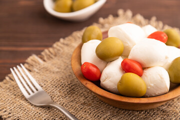 White Mozzarella cheese, with tomatoes and olives on brown wooden, linen textile, side view, close up, selective focus.