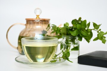 A transparent cup of fresh mint tea, next to an elegant teapot and a bunch of mint leaves in water.